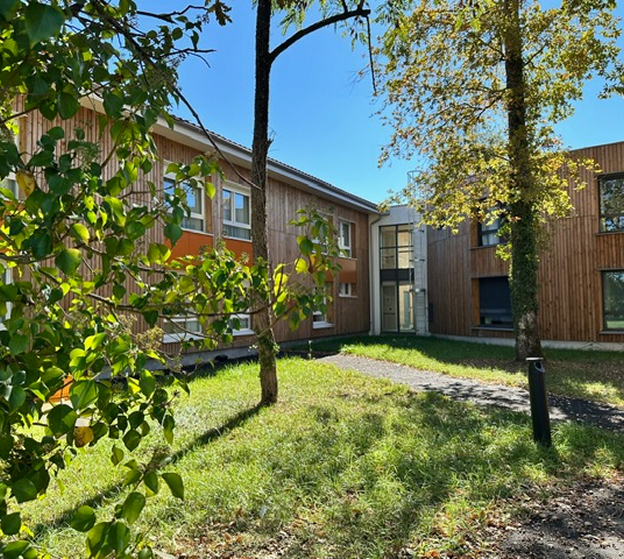 Deux bâtiments en bois, une allée et des arbres verdoyants sous un ciel bleu clair à Rochefort en Charente-Maritime 17